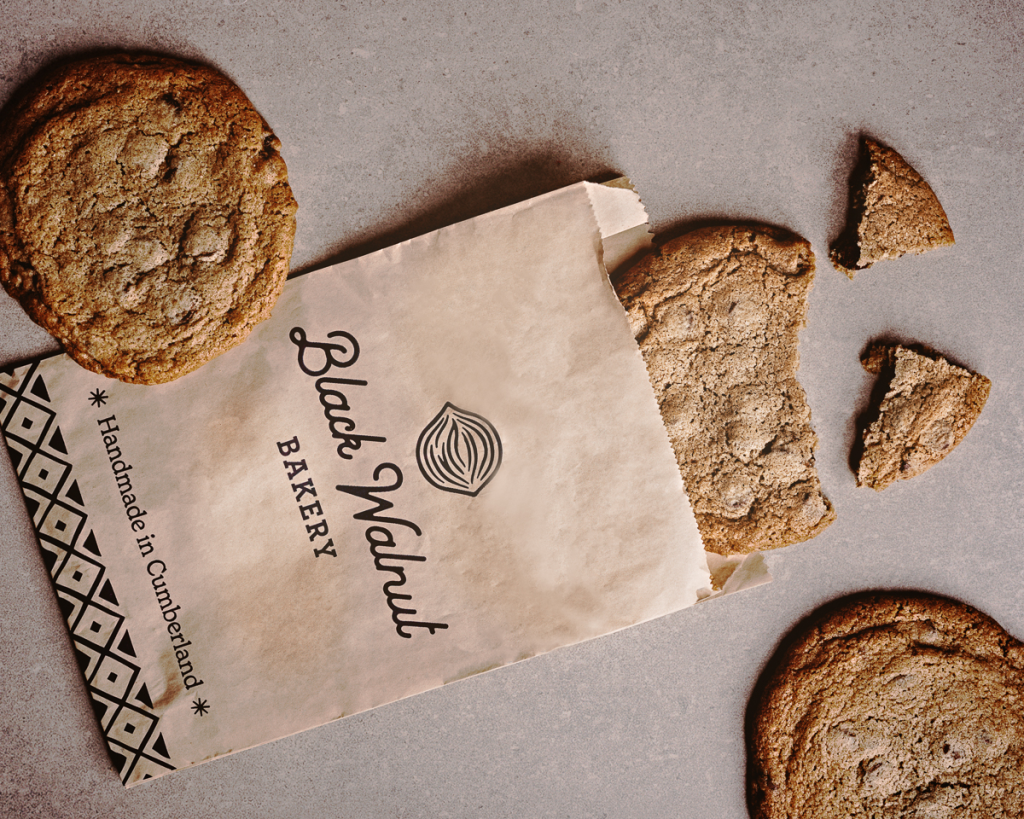 Three cookies laid out on a concrete counter, with a brown paper bag from Black Walnut Bakery. One of the cookies is broken into 3 pieces. Creative product photography by Ottawa commercial photographer Seb Duper.