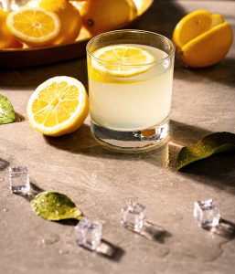 A glass of cold, fresh lemonade, with a slice of lemon floating on top. A lemon sliced in half leans against the left side of the glass, brightly lit by the sun. In the foreground, we can see melting ice cubes and mint leaves with water drops on them. Creative food & drink photography by Ottawa commercial photographer Seb Duper.