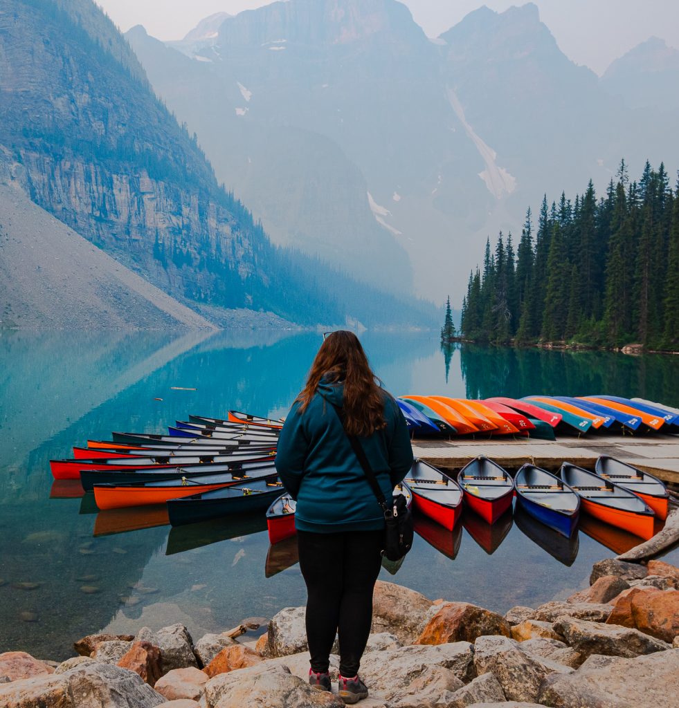 A woman with her back to the camera stares out at Moraine Lake, with colourful canoes in the foreground.