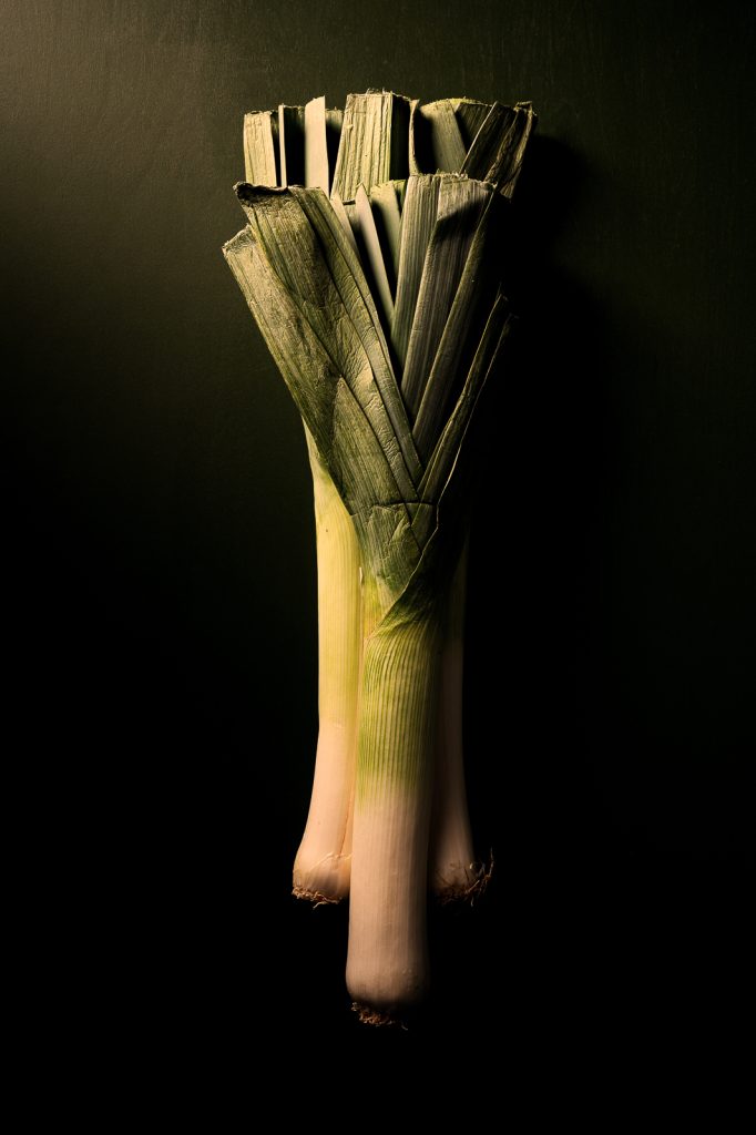 Three bunches of leeks on a dark background. Soft, warm light coming from the top left corner. Product photography by Seb Duper.