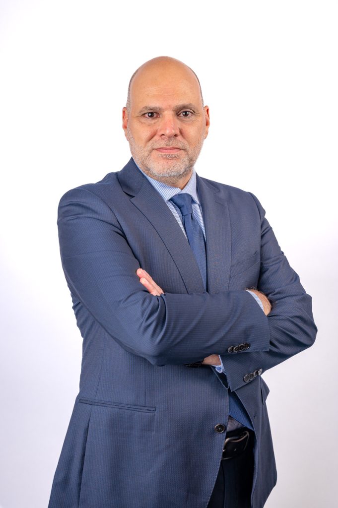 Headshot of a man wearing a blue blazer, blue shirt and blue tie, against a white background. His arms are crossed over his chest.