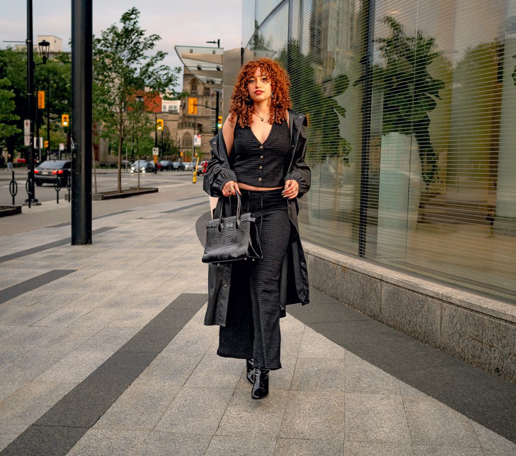 A young woman dressed in black pants, a black shirt and black coat, holding a black handbag as she walks along an Ottawa street.