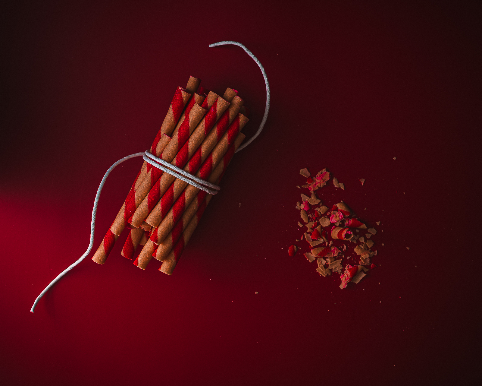 On the left, a bunch of red and white wafer rolls tied together with white cord, and wafer crumbs on the right. The background is dark red, and there's some light coming in from the bottom left.