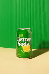 A can of soda resting on a butter-yellow tabletop, and set in front of a bright green, textured background.